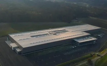 Eine Luftaufnahme eines großen Fabrikgebäudes mit Solarpanelen auf dem Dach, umgeben von einem Parkplatz und Grünflächen.