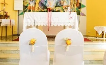 Zwei mit weißem Stoff bezogene Stühle, geschmückt mit gelben Blumen, stehen vor einem Altar in einer Kirche. Hinter ihnen steht ein weißer Altartisch, der mit Blumen und Kerzen geschmückt ist. Ein großes Wandbild befindet sich hinter dem Altar.