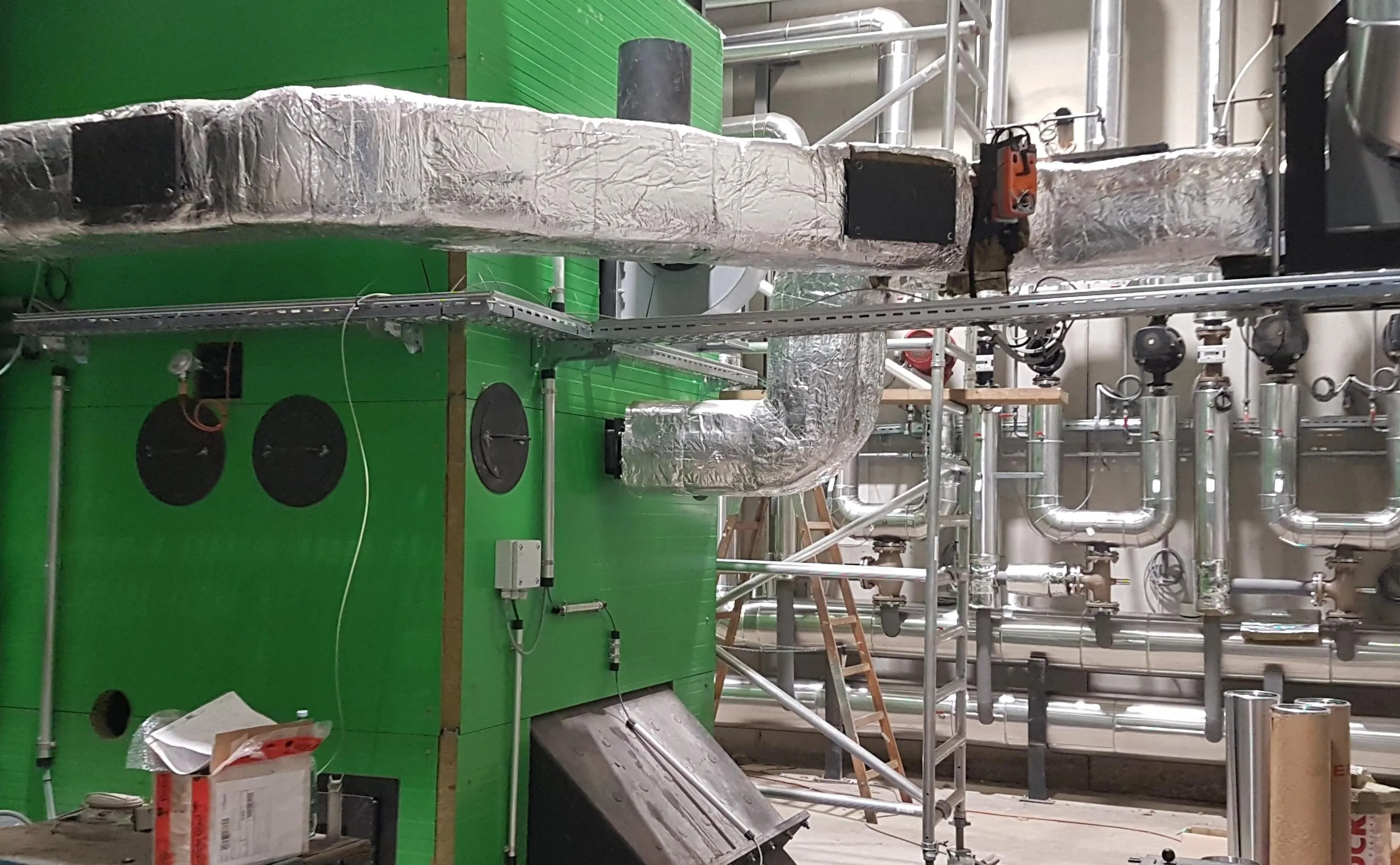 Ein Innenraumbild einer grünen Struktur mit einem angeschlossenen metallischen Rohrsystem. Ein großer Metallrohr ist in Aluminiumfolie eingewickelt.