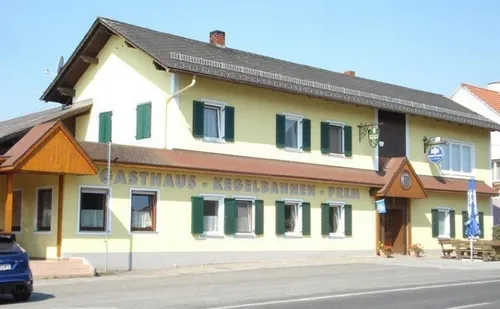 Ein gelbes Gebäude mit grünen Fensterläden hat ein Schild, das Gasthaus Kegelbahnen Prem besagt. Ein blaues Auto ist davor geparkt.