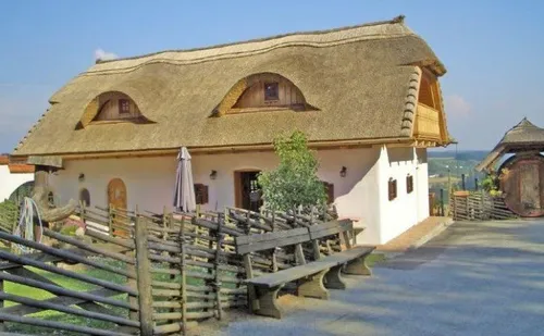 Ein Haus mit einem Strohdach, Holzwänden und einer Veranda mit einer Bank und einem geschlossenen Regenschirm. Das Haus hat Bogenfenster.