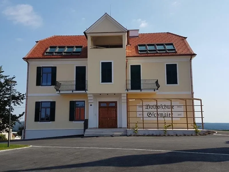 Ein gelbes Haus mit rotem Dach und braunen Türen, mit mehreren Fenstern, Balkonen und einem Garten davor.