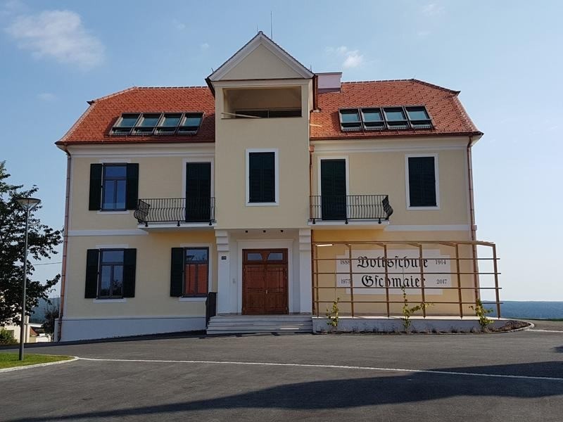 Ein gelbes Haus mit rotem Dach und braunen Türen, mit mehreren Fenstern, Balkonen und einem Garten davor.