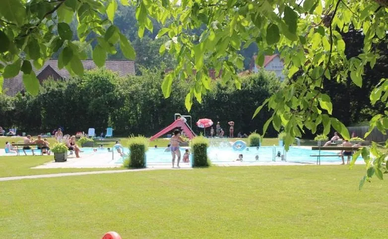 Ein großer Swimmingpool ist an einem sonnigen Tag überfüllt mit Menschen. Es gibt mehrere Menschen, die am Poolrand sitzen, und andere schwimmen. Es gibt eine rote Rutsche in der Nähe des Pools. Es gibt Pflanzen und Bäume um den Pool herum.