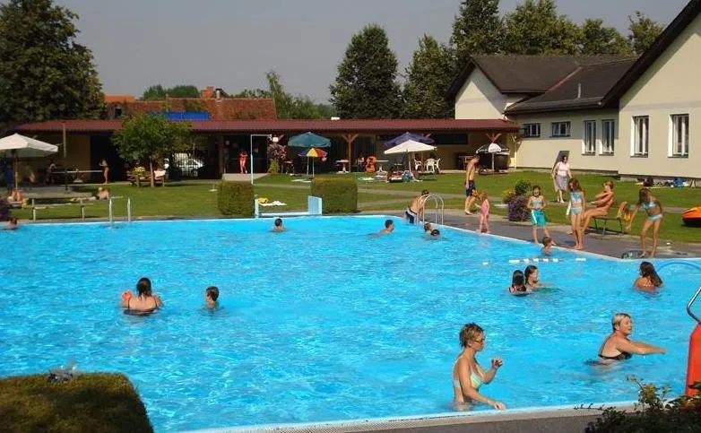 Menschen schwimmen in einem großen Pool, umgeben von einem Gebäude, Bäumen und einer Rasenfläche. Einige Personen entspannen sich unter Sonnenschirmen auf dem Rasen.