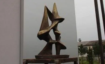 Eine Bronzeskulptur abstrakter Figuren steht auf einer Holzplattform. Die Skulptur besteht aus drei miteinander verbundenen Formen, die möglicherweise eine Familie oder Einheit darstellen. Der Ort ist ein Raum mit weißen Wänden und einem bewölkten Himmel im Hintergrund.