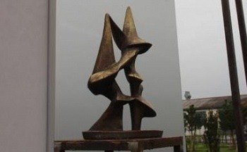 Eine Bronzeskulptur abstrakter Figuren steht auf einer Holzplattform. Die Skulptur besteht aus drei miteinander verbundenen Formen, die möglicherweise eine Familie oder Einheit darstellen. Der Ort ist ein Raum mit weißen Wänden und einem bewölkten Himmel im Hintergrund.
