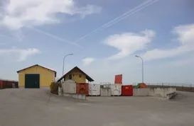 Ein leerer Platz mit zwei Gebäuden, eines gelb und eines braun, hat Straßenlaternen und eine Betonbarriere. Der Himmel ist blau mit verstreuten Wolken.