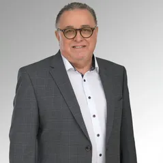 Ein Mann in einem grauen Blazer und einem weißen Hemd mit schwarzen Knöpfen. Er lächelt und steht vor einem grauen Hintergrund.