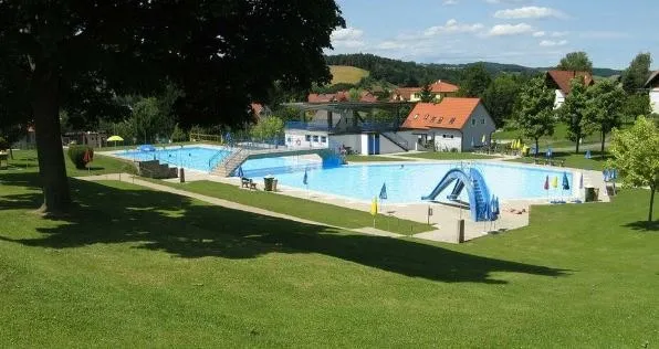 Ein großer Außenpool mit Sprungbrett und Rutsche, umgeben von üppigem grünem Rasen, Bäumen und Häusern in der Ferne.