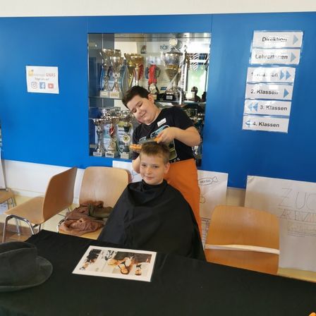 Ein Junge mit kurzen Haaren sitzt in einem Stuhl und lässt sich die Haare schneiden. Dahinter ist eine blaue Wand mit ausgestellten Trophäen.