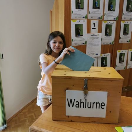 Ein junges Mädchen gibt ihre Stimme in eine Holzurne ab, die mit 'Wahlurne' beschriftet ist, vor einer Wand mit verschiedenen Plakaten.