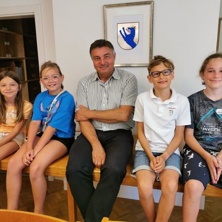 Ein Mann und vier Kinder sitzen auf einer Bank. Der Mann lächelt und hat die Arme verschränkt. Drei der Kinder tragen eine Brille. Hinter ihnen ist eine weiße Wand mit einem Bilderrahmen.