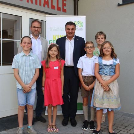 Eine Gruppe von Erwachsenen und Kindern steht vor einem Gebäude mit einem Banner und einem Schild, auf dem 'Kindergarten' steht. Sie lächeln und posieren für ein Foto.