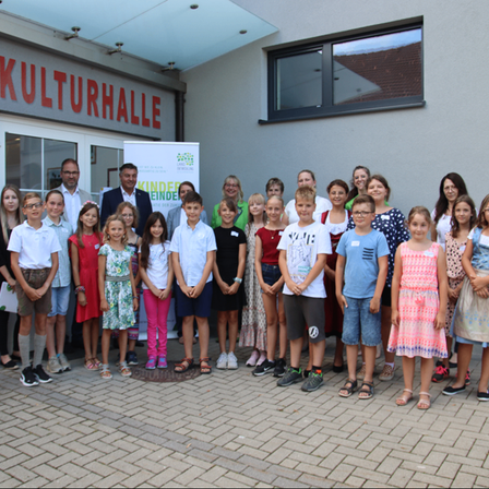 Eine Gruppe von Kindern und Erwachsenen steht vor dem Gebäude der Kulturhalle und lächelt für ein Foto.