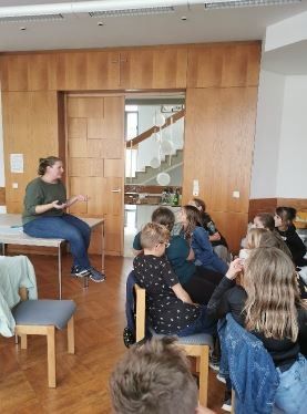 Eine Frau sitzt auf einer Bank in einem Klassenzimmer und spricht zu einer Gruppe sitzender Schüler. Im Hintergrund befinden sich Holzwände und eine Treppe.