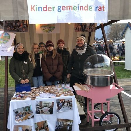 Eine Gruppe von Menschen steht hinter einem Tisch voller Donuts. Daneben befindet sich eine Zuckerwattemaschine. Es gibt Plakate an der Wand. Der Banner lautet Kinder Gemeinde Rat.