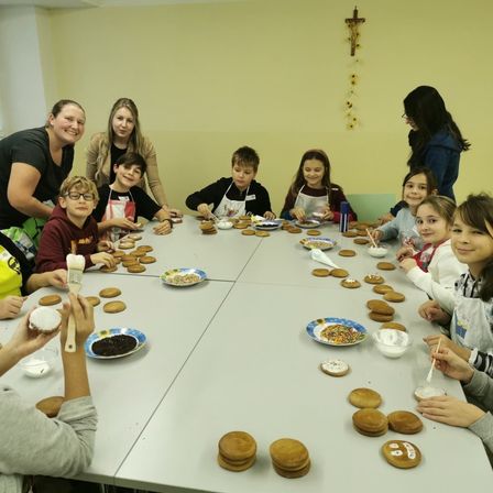 Eine Gruppe von Kindern und Erwachsenen verziert Kekse an einem Tisch, einige mit Streuseln und Zuckerguss. Eine Frau hält einen Pinsel, während andere Zuckerbeutel benutzen. Ein Kreuz hängt an der Wand.