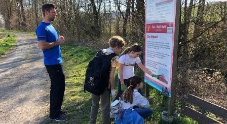 Ein Mann und drei Kinder stehen neben einem Schild auf einem Grasweg und betrachten es genau.