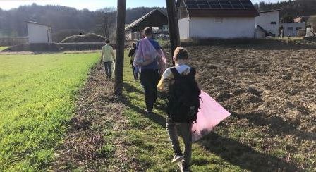 Eine Gruppe von Menschen geht entlang eines grasbewachsenen Weges in der Nähe eines Feldes. Einer trägt einen Rucksack und eine rosa Tasche. Gebäude und Bäume sind im Hintergrund.