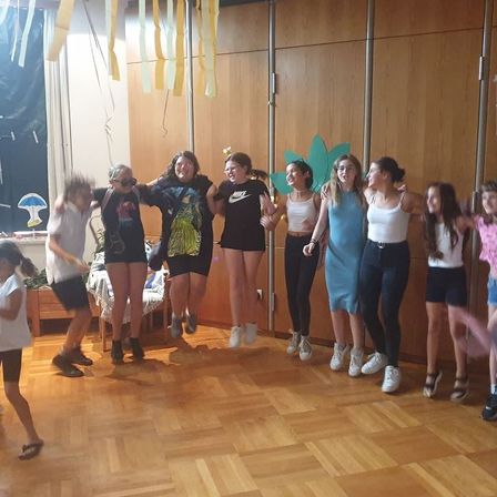 Eine Gruppe von Frauen und Kindern steht in einem Raum, einige springen und halten sich an den Händen. Eine Pflanze ist an der Wand.