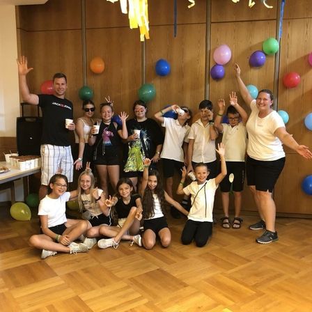 Eine Gruppe von Kindern und Erwachsenen posiert für ein Foto bei einer Veranstaltung. Sie alle lächeln und halten Tassen. Der Raum ist mit Ballons und Girlanden geschmückt.