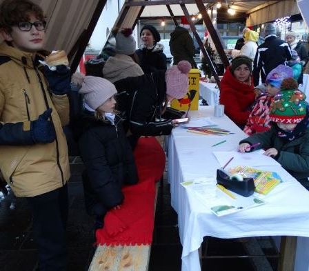 Mehrere Kinder, einige mit Mützen, sitzen an einem Tisch und zeichnen. Ein Junge steht neben ihnen und hält ein Sandwich. Andere Leute sitzen und stehen im Hintergrund.