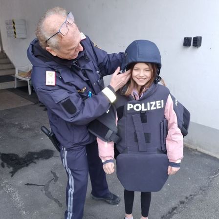 Ein Polizist in Uniform hilft einem jungen Mädchen, eine kugelsichere Weste anzuziehen. Sie stehen in einem Parkplatz und lächeln.