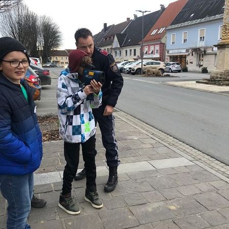 Ein Polizist steht mit zwei Kindern auf dem Bürgersteig, eines hält eine Kamera. Hinter ihnen sind geparkte Autos und eine Straßenlaterne.