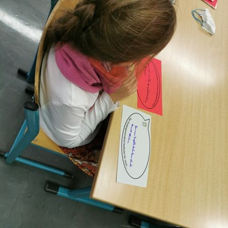 Ein Kind sitzt an einem Schreibtisch und schaut auf ein Papier mit Sprechblasen. Das Papier hat fremde Schriftzeichen. Der Schreibtisch ist braun und der Stuhl blau.