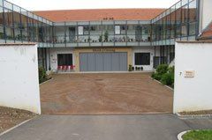 Ein modernes Gebäude mit rotem Dach und Glaswänden steht vor einem leeren Parkplatz. Der Eingang ist durch zwei weiße Wände gekennzeichnet, mit einer grauen Garagentür in der Mitte.