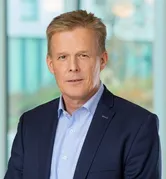 Ein Mann mit kurzen blonden Haaren, der einen dunkelblauen Anzug und ein hellblaues Hemd trägt, steht vor einer Glaswand.