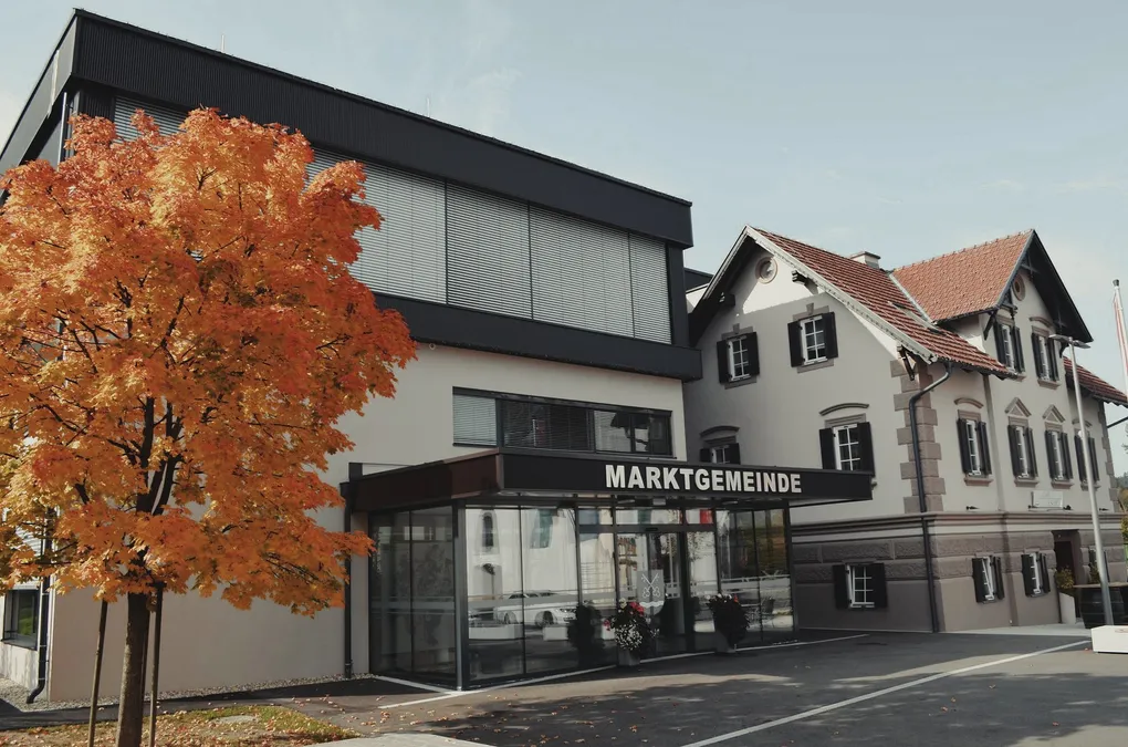 Abschnittsbild: Ein modernes Gebäude mit schwarzem Dach und Glaswänden, benannt 'MARKTGEMEIND', steht vor einem Baum mit orangefarbenen Blättern.