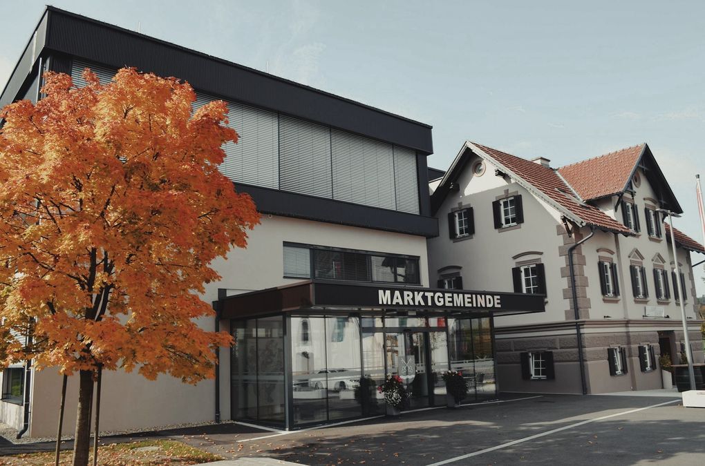  Abschnitt Bild: Ein modernes Gebäude mit schwarzem Dach und Glaswänden, benannt 'MARKTGEMEIND', steht vor einem Baum mit orangefarbenen Blättern.