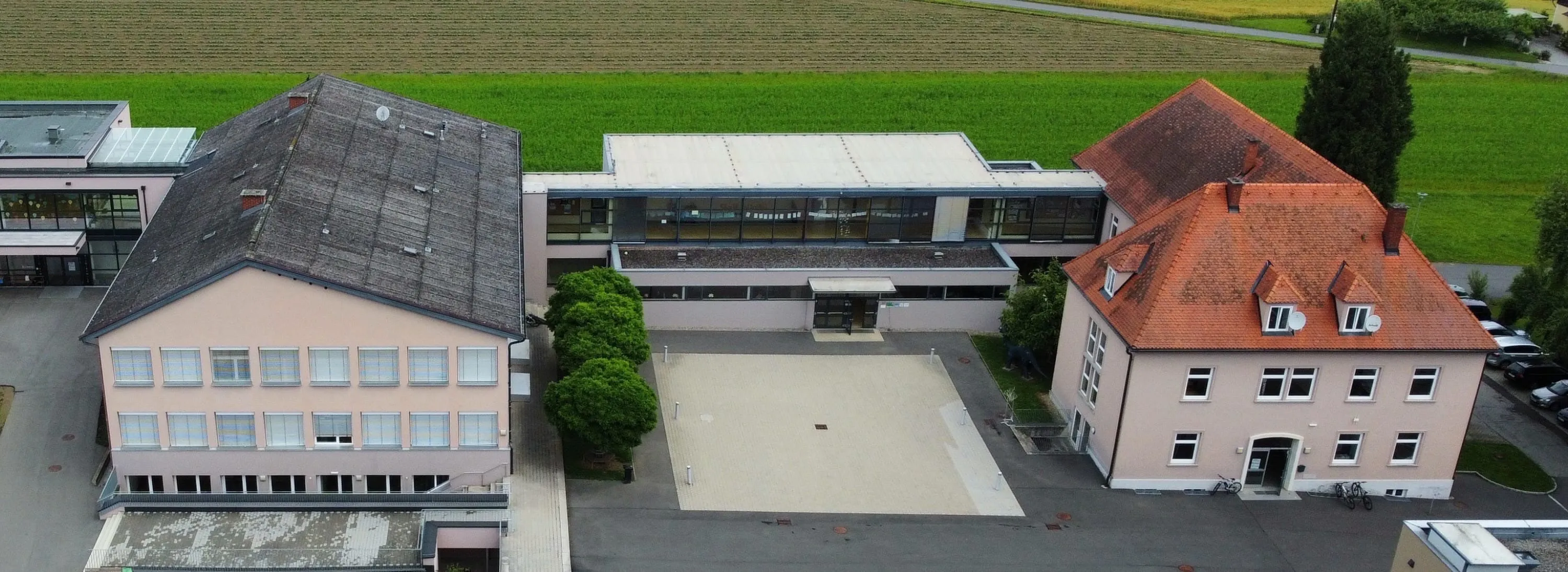 Luftaufnahme eines Gebäudes mit großen Fenstern und einem gepflasterten Innenhof, umgeben von einem grünen Feld und kleinen Bäumen.