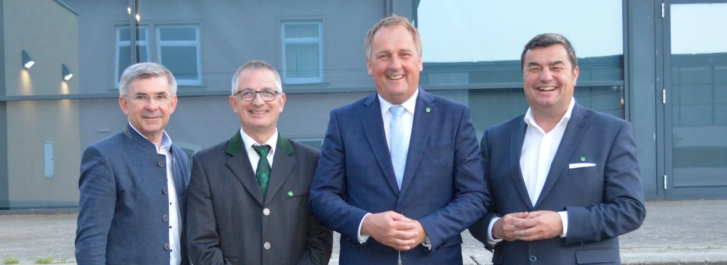 Drei Männer lächeln vor einem Gebäude. Der Mann in der Mitte trägt einen blauen Anzug und eine Krawatte. Der Mann auf der linken Seite trägt eine grüne Krawatte und eine grüne Schärpe. Der Mann auf der rechten Seite trägt eine Uhr und einen blauen Anzug.
