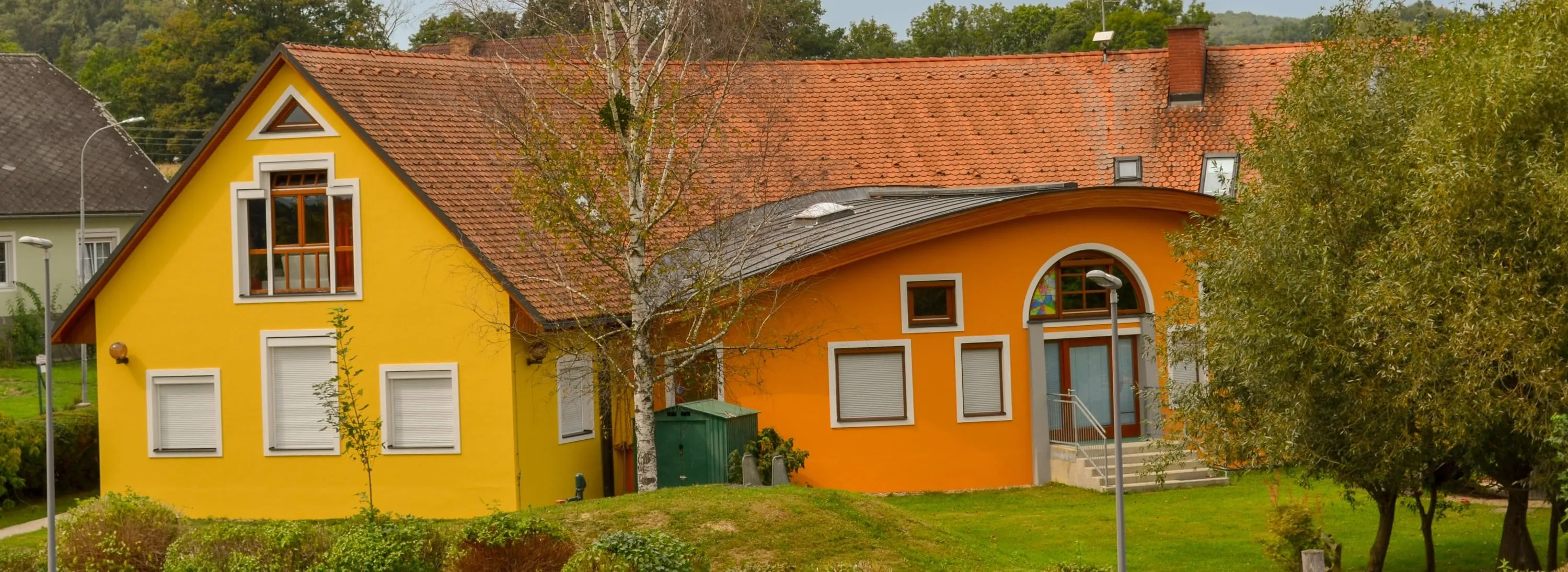 Ein leuchtend oranges Haus mit einem roten Dach, weißen Fensterrahmen und drei Fenstern. Ein kleiner grüner Schuppen steht im Garten, mit einem Baum im Vordergrund.