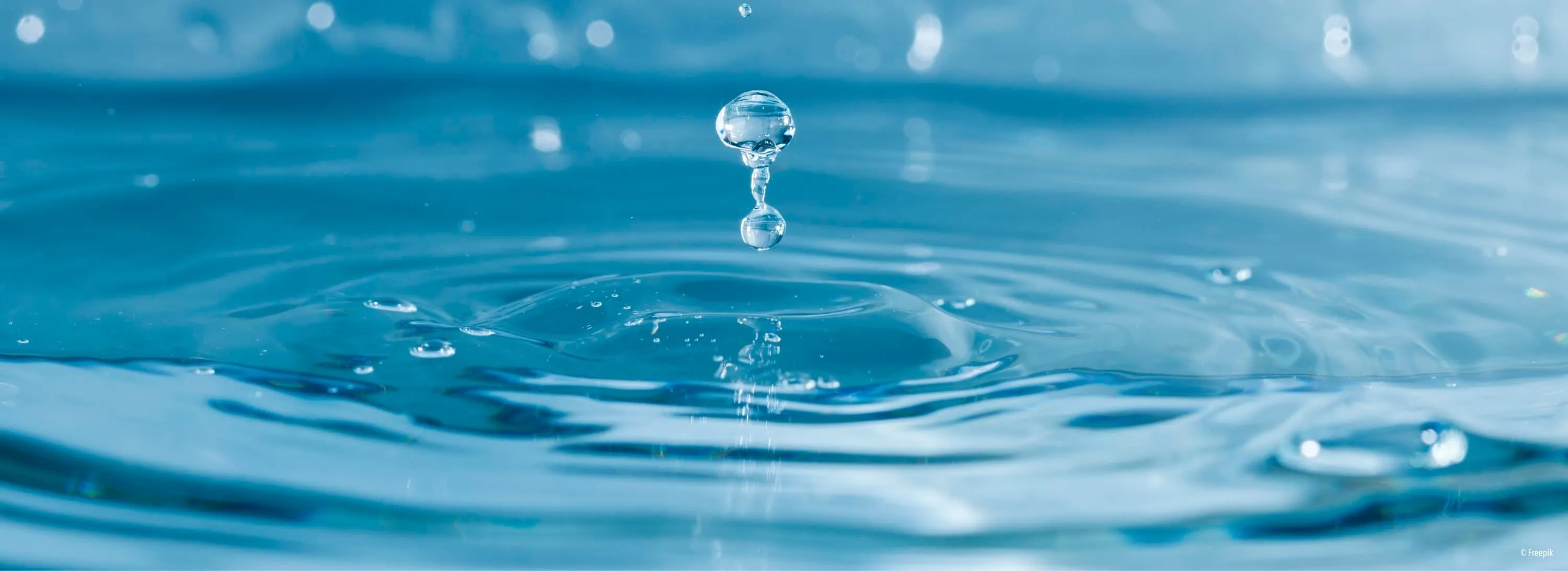 Ein Wassertropfen fällt in einen klaren blauen Pool und erzeugt Wellen. Das Wasser ist rein, mit kleinen Blasen sichtbar.