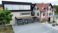 Luftaufnahme eines Gebäudes mit dem Text 'Marktgemeinde' auf einem Vordach, zwei Topfpflanzen und einem Haus mit rotem Dach.