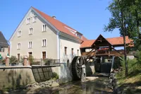 Ein historisches Gebäude mit rotem Dach und vielen Fenstern, das ein großes Wasserrad und eine hölzerne Brücke mit einem Balkon mit roten Blumen aufweist.