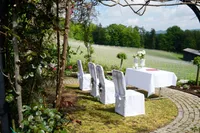 Eine Außenveranstaltung für eine Hochzeit mit in einer Reihe angeordneten Stühlen und einem in Weiß gedeckten Tisch. Im Hintergrund befindet sich ein Weinberg mit üppigem Grün und Bergen.