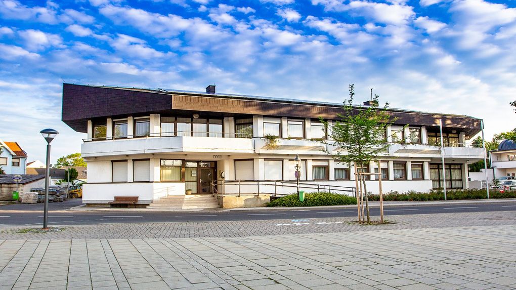  Abschnitt Bild: Ein weißes Gebäude mit vielen Fenstern und einem braunen Dach steht am Straßenrand.