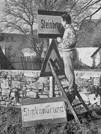 Ein Mann steht auf einer Leiter und berührt ein Schild mit dem Wort 'Steinbro', mit einem weiteren Schild und einem Warndreieck neben ihm, vor einer Steinmauer im Hintergrund.