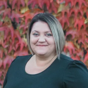 Eine Frau mit kurzen blonden Haaren lächelt vor einer roten Ahornblattwand.