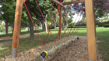 Eine Reihe roter Seile ist zwischen Holzbalken in einem Park aufgehängt und bildet einen Hängeweg. Der Weg besteht aus Seilen mit gelben und grauen Abdeckungen und ist von grünem Gras und Bäumen umgeben.