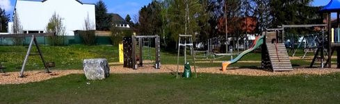 Ein Spielplatz im Freien mit Rutsche, Schaukeln und Klettergeräten in einem Grasbereich mit Bäumen im Hintergrund.