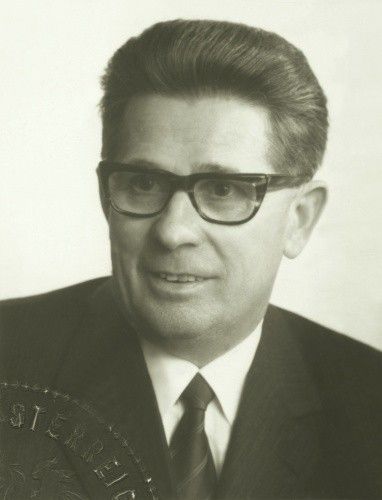 Schwarz-Weiß-Foto eines Mannes in formeller Kleidung, der eine Brille und eine Krawatte trägt. Er scheint sich in einem professionellen Umfeld zu befinden, und ein Medaillon ist an seiner Reversnadel sichtbar.