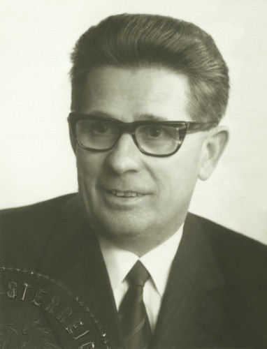 Schwarz-Weiß-Foto eines Mannes in formeller Kleidung, der eine Brille und eine Krawatte trägt. Er scheint sich in einem professionellen Umfeld zu befinden, und ein Medaillon ist an seiner Reversnadel sichtbar.