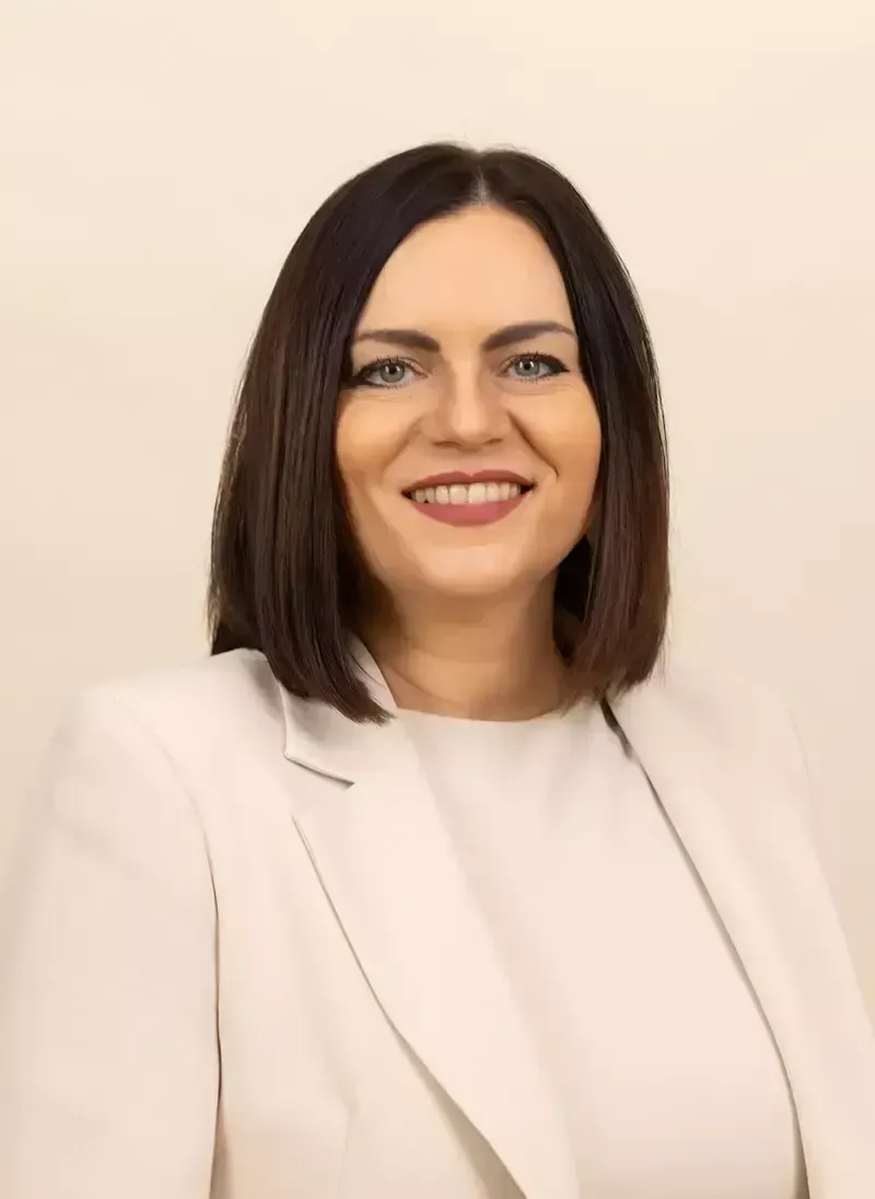 Eine Frau mit kurzen braunen Haaren und blauen Augen lächelt und posiert für ein Foto. Sie trägt einen weißen Blazer und ein weißes Hemd.