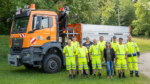 Eine Gruppe von Personen in gelben Sicherheitsanzügen posiert vor einem orangefarbenen Lastwagen mit Schneepflug-Aufbau.